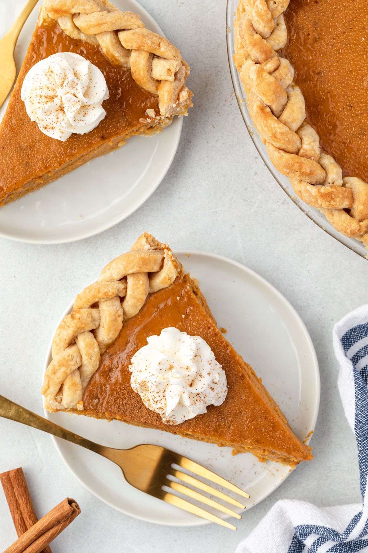 Slice of pumpkin pie with a braided crust and a dollop of whipped cream with a dusting of pumpkin spice on a plate with a gold fork.