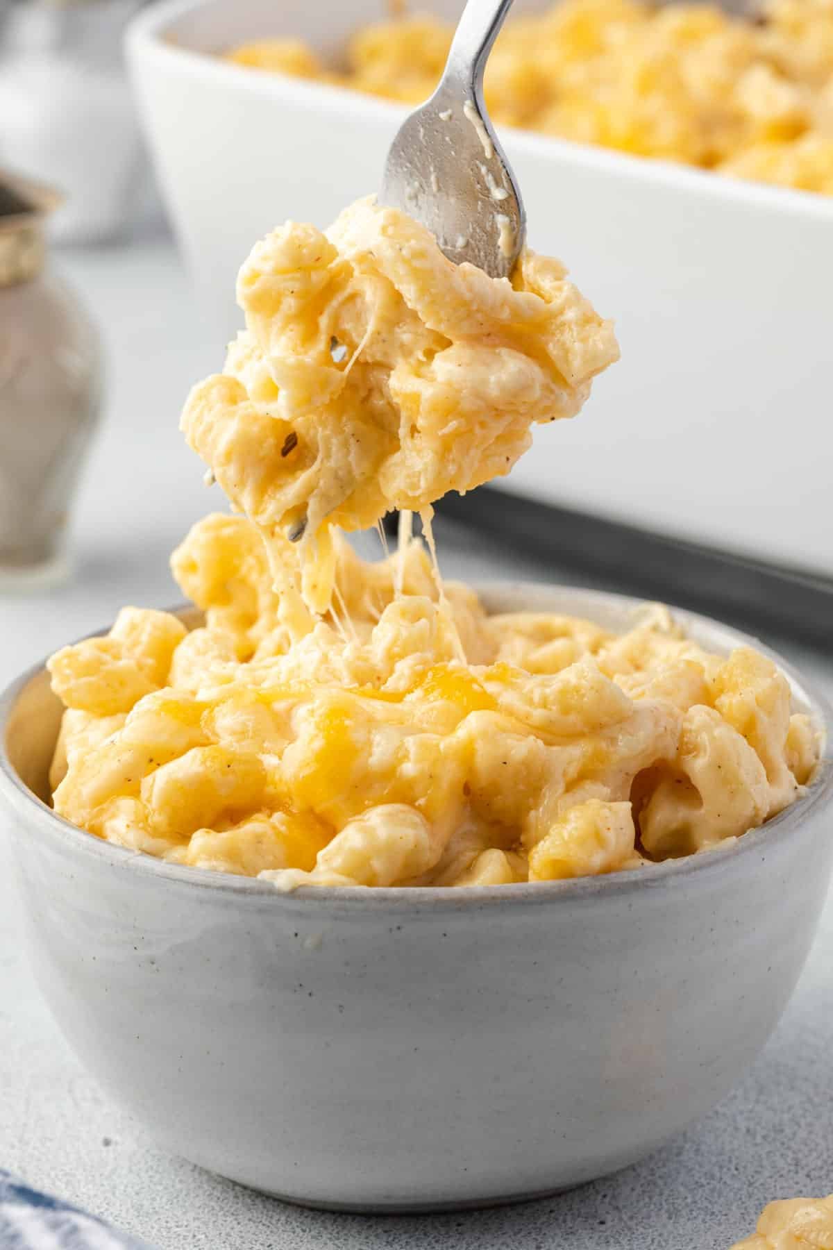 A fork with mac and cheese held over a bowl of macaroni with a cheese pull.