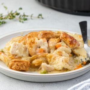 A serving of pot pie filling topped with puff pastry on a plate, with sprig of thyme and pie plate with whole pot pie.