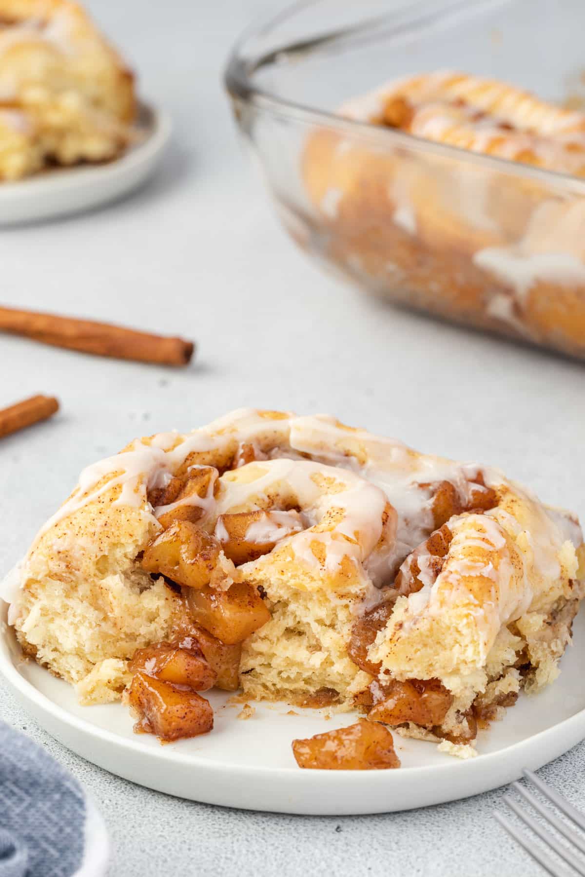 Soft cinnamon roll on a plate with apple pie filling spilling out, and the pan of rolls in background.