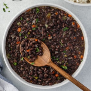 Overhead of cuban-style black beans in a white bowl with a wooden spoon scooping some beans.