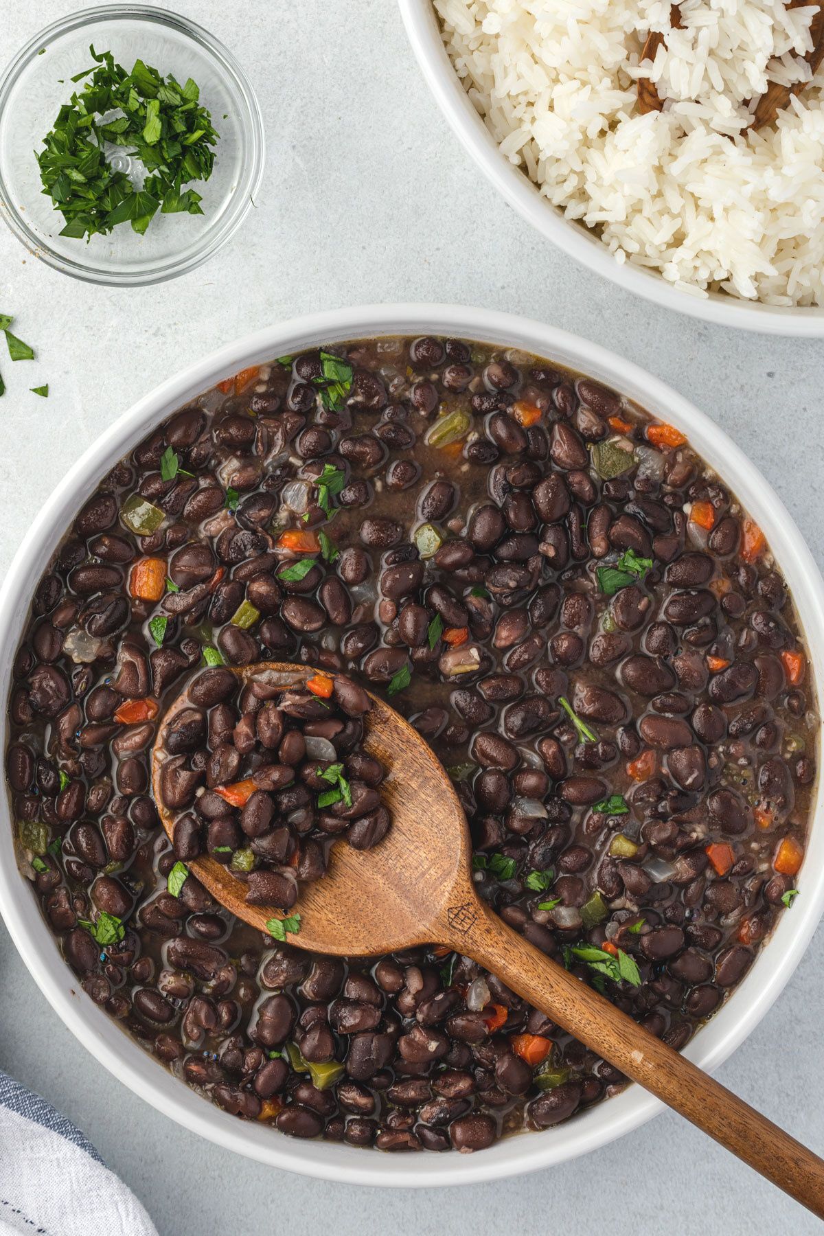 Overhead of cuban-style black beans in a white bowl with a wooden spoon scooping some beans. cilantro and white rice in corners..