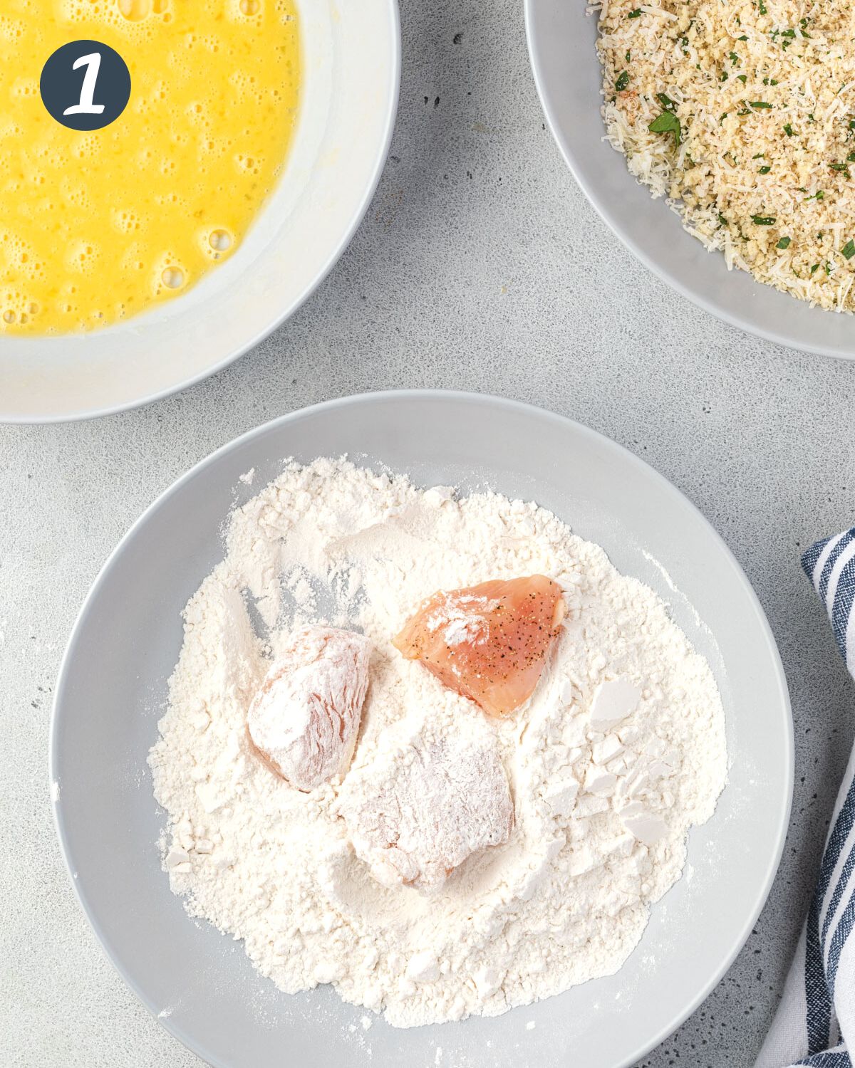 A shallow grey bowl of flour with 3 pieces of chicken, 2 coated in flour and one plain.