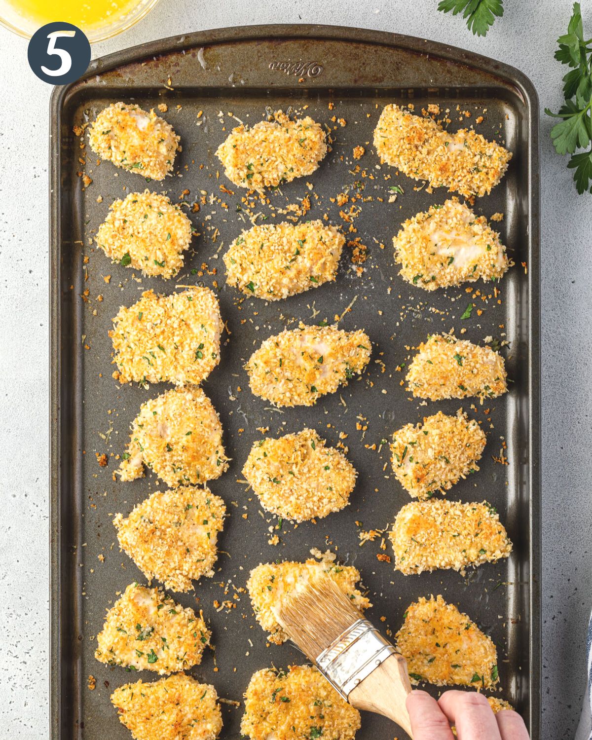 Brushing butter with a pastry brush onto a baking sheet of breaded chicken pieces.