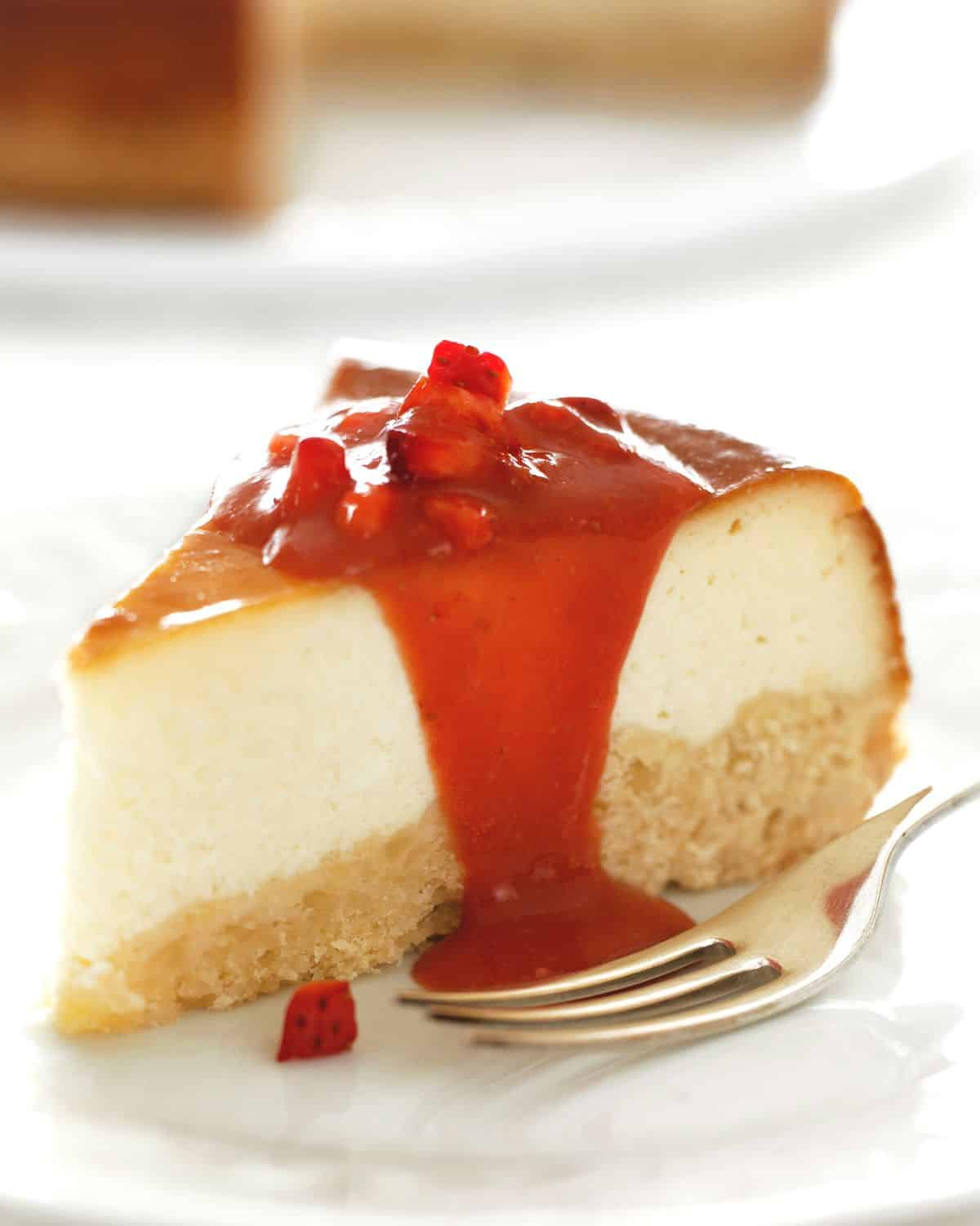 A slice of cheesecake on a plate with strawberry fruit sauce dropping down.