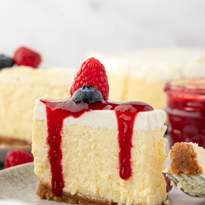 Slice of cheesecake topped with raspberry sauce, a raspberry, and a blueberry on a plate.