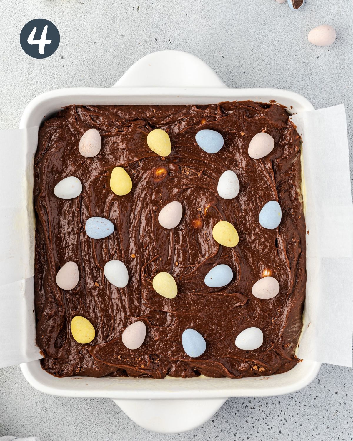 Overhead of a square baking dish with white handles, filled with brownie mix topped with mini eggs.