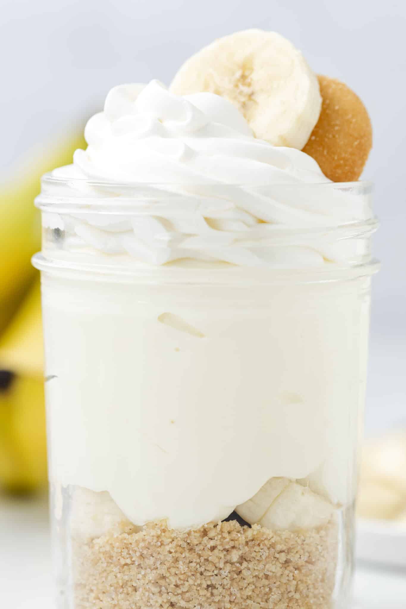 A mason jar with graham cracker crumbs in the bottom, filled with cheesecake filling, and topped with whipped cream, banana slice, and vanilla wafer.