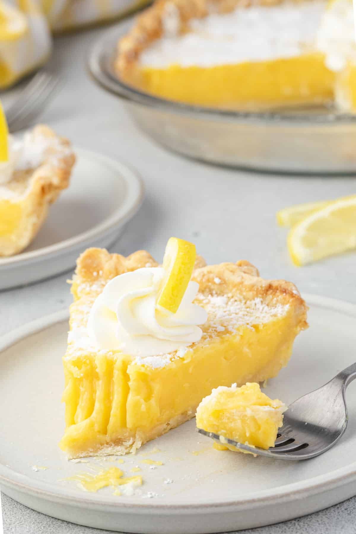 Slice of lemon pie on a plate with a bite on a fork, and the cut whole pie in the background.
