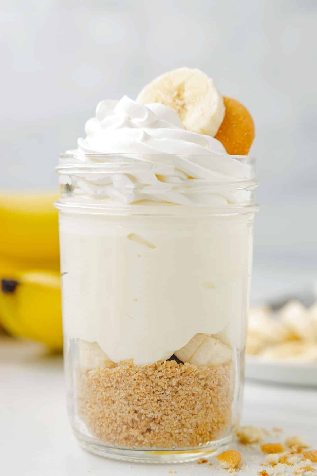 Single jar of no bake cheesecake, showing off the layers, with crumbs in front of the jar and bananas in background.