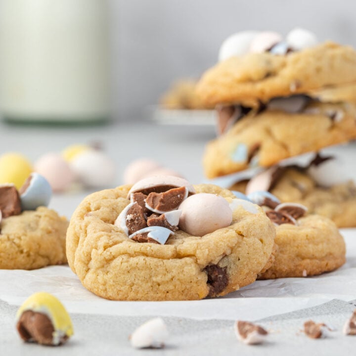 Side view of 3 mini egg cookies shingled on white parchment with broken candy pieces around, a stack of cookies and a jar of milk in background.