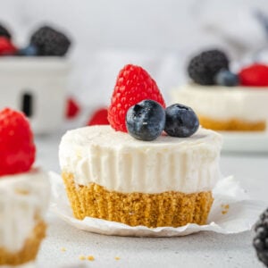 An individual mini cheesecake with paper liner peeled back, berries on top and more mini cheesecakes and a basket of berrries in background.