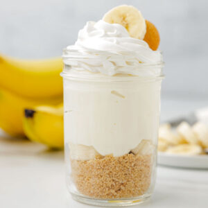 Single jar filled with cookie crumb filling, banana slices, cheesecake, and topped with a piped whipped cream, a banana slice, and vanilla wafer cookie.