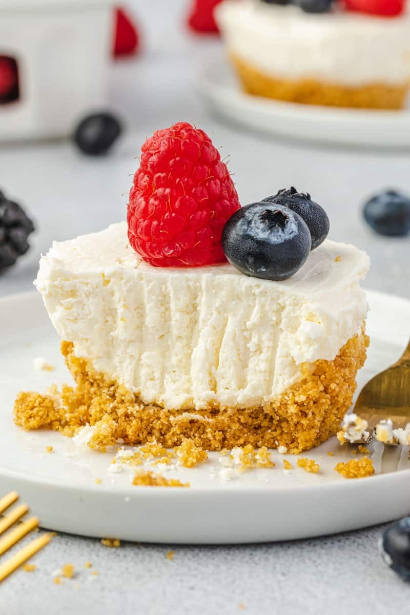 A mini cheesecake topped with berries on a plate with the bites missing and fork tine marks in cheesecake.