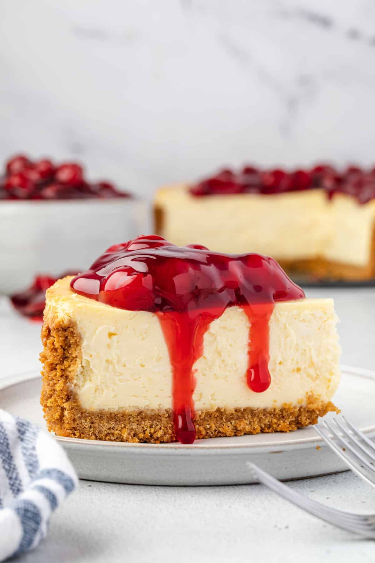 Slice of classic philadelphia cheesecake with graham cracker crust, topped with cherry pie filling that is dripping down side of cheesecake.