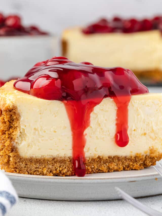Classic cheesecake slice on a plate with cherry pie filling on top, bowl of cherries and whole cheesecake in background.