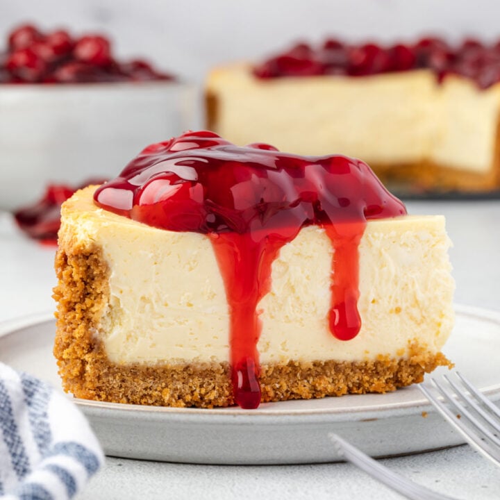 Classic cheesecake slice on a plate with cherry pie filling on top, bowl of cherries and whole cheesecake in background.