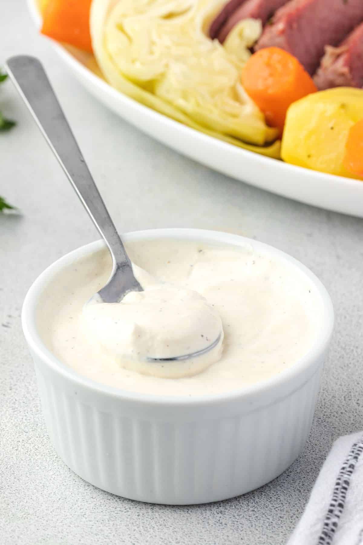 White ramekin filled with mustard sauce, spoon in sauce and platter of corned beef and vegetables in corner.
