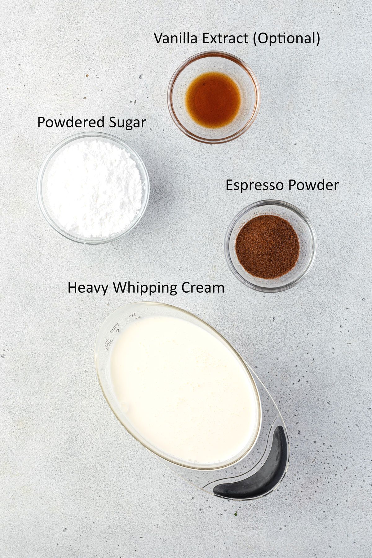 Overhead showing measuring cup of whipped cream, and individual bowls of powdered sugar, espresso powder, and vanilla extract.