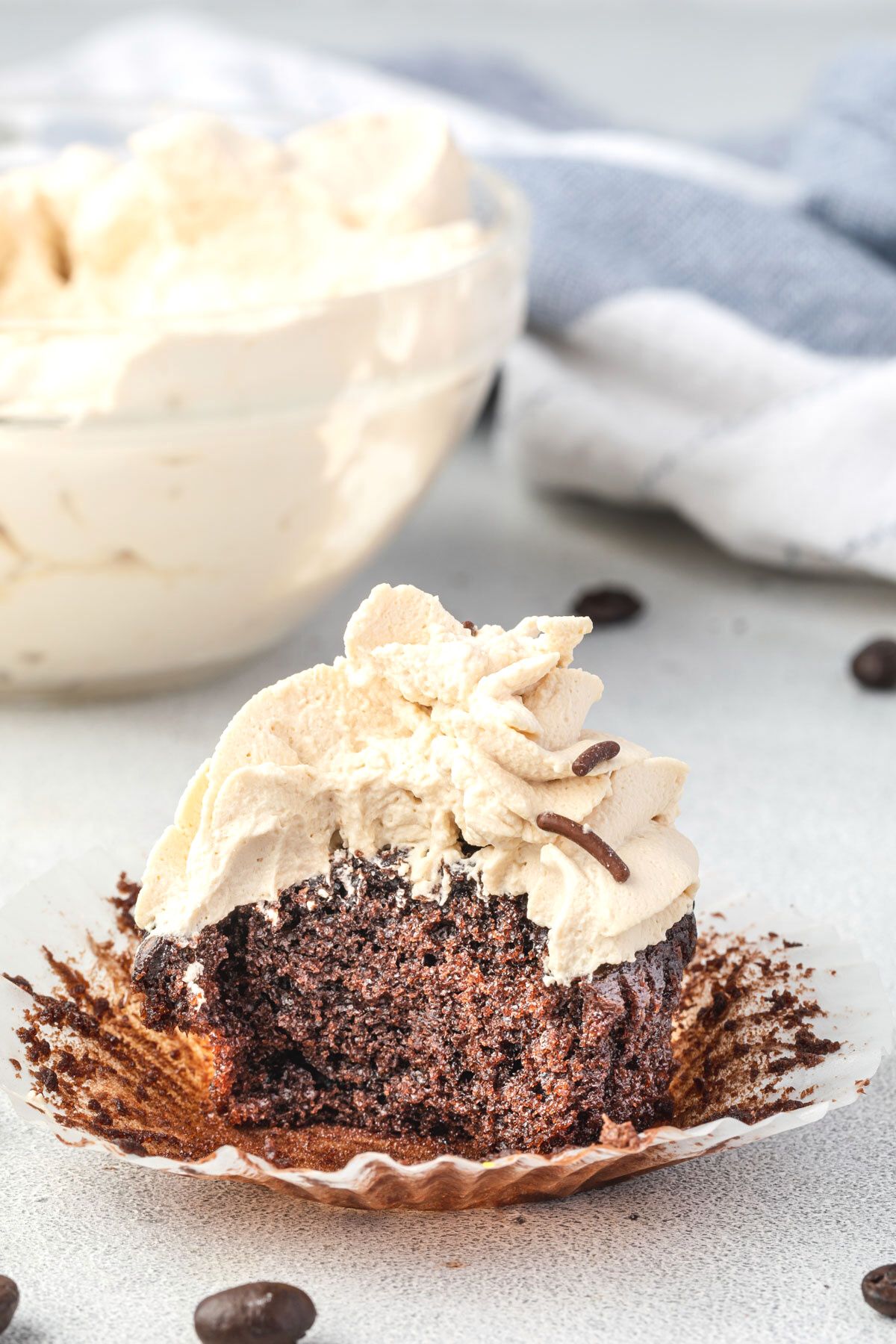 A chocolate cupcake with the wrapper peeled down, topped with espresso whipped cream - a bowl of cream and coffee beans in background.