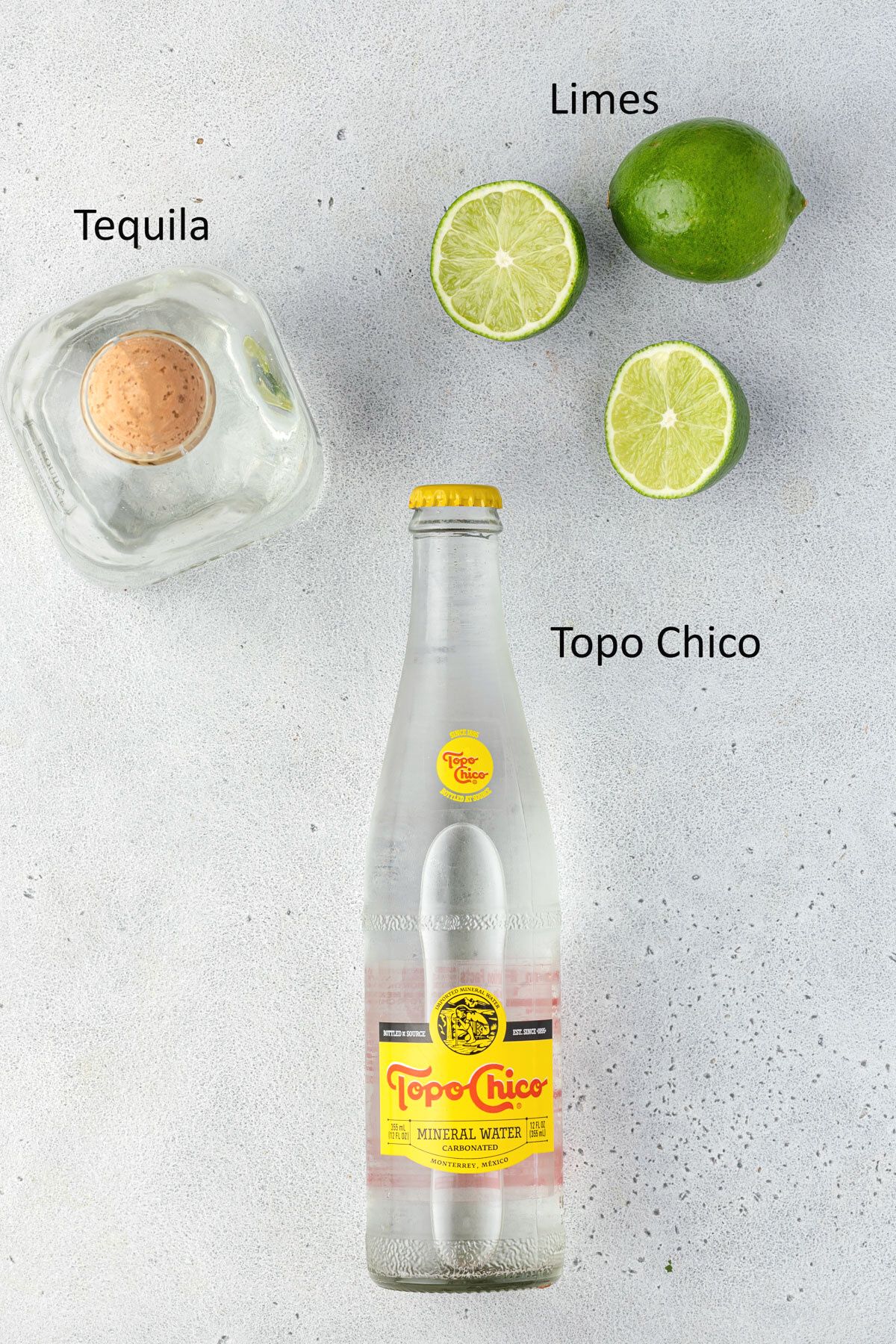 Overhead of bottle of tequila, Topo Chico, and limes on a cement surface.