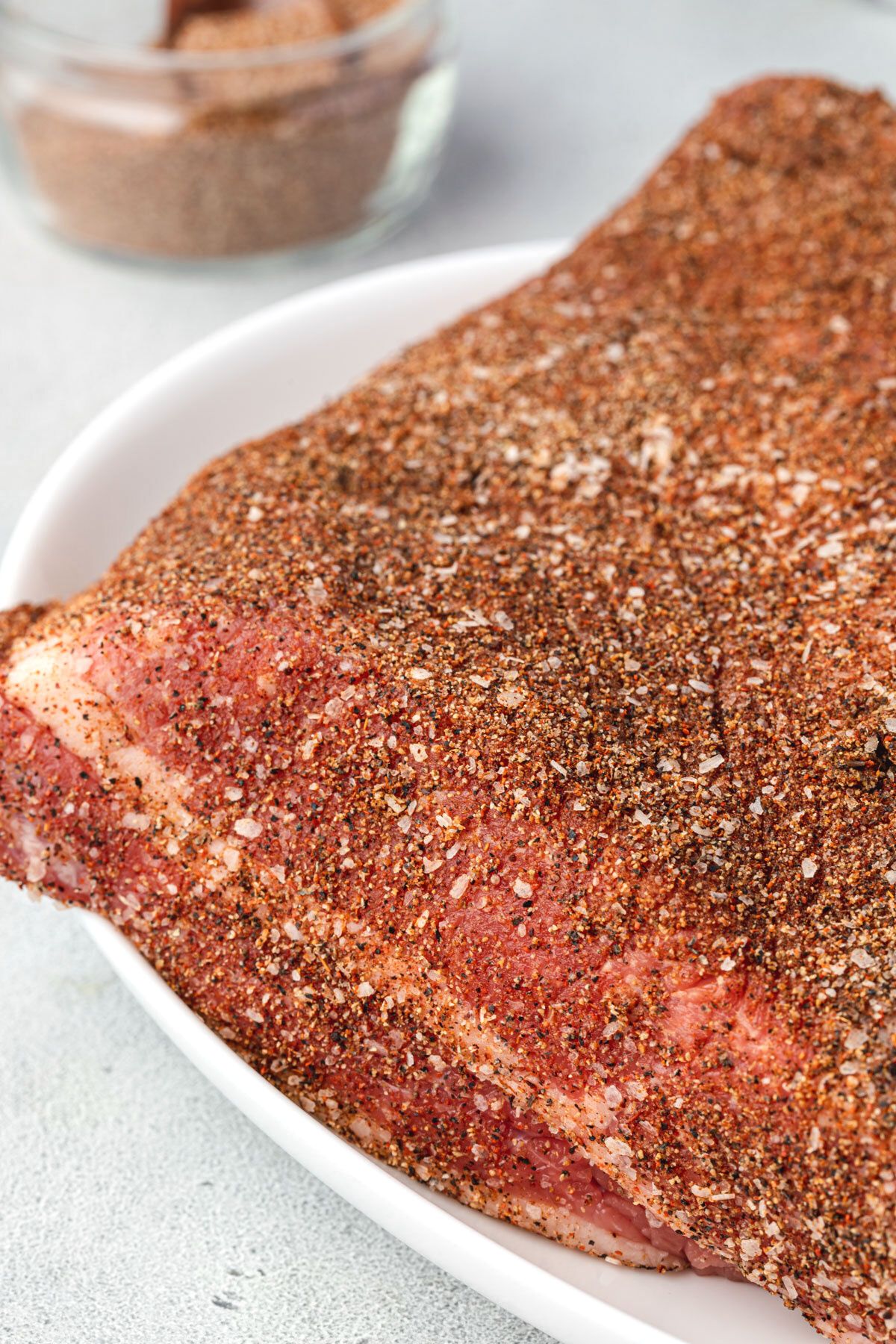 Close up of raw brisket coated with spice rub on a white platter with bowl of rub in background.