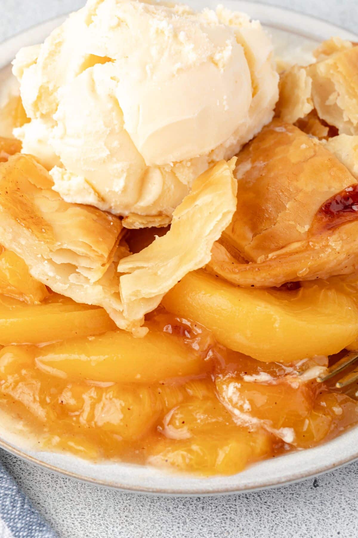 Close up of peach cobbler on a plate, showing slices of peaches, flaky crust topping, and melty ice cream.