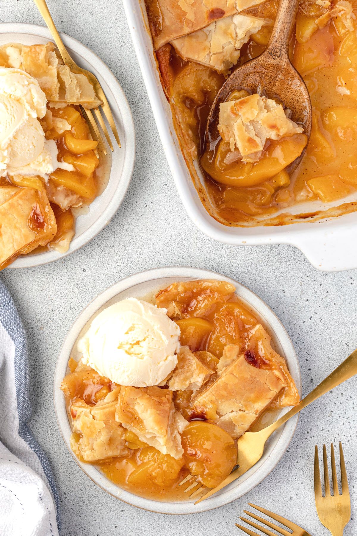 Overhead of 2 plates of peach cobbler with ice cream on top and gold forks, and corner showing of the pan of cobbler.