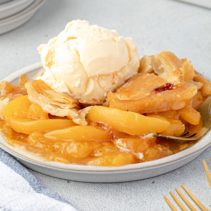 Plate of peach cobbler with juicy peaches and flaky pie crust topped with a scoop of vanilla ice cream.