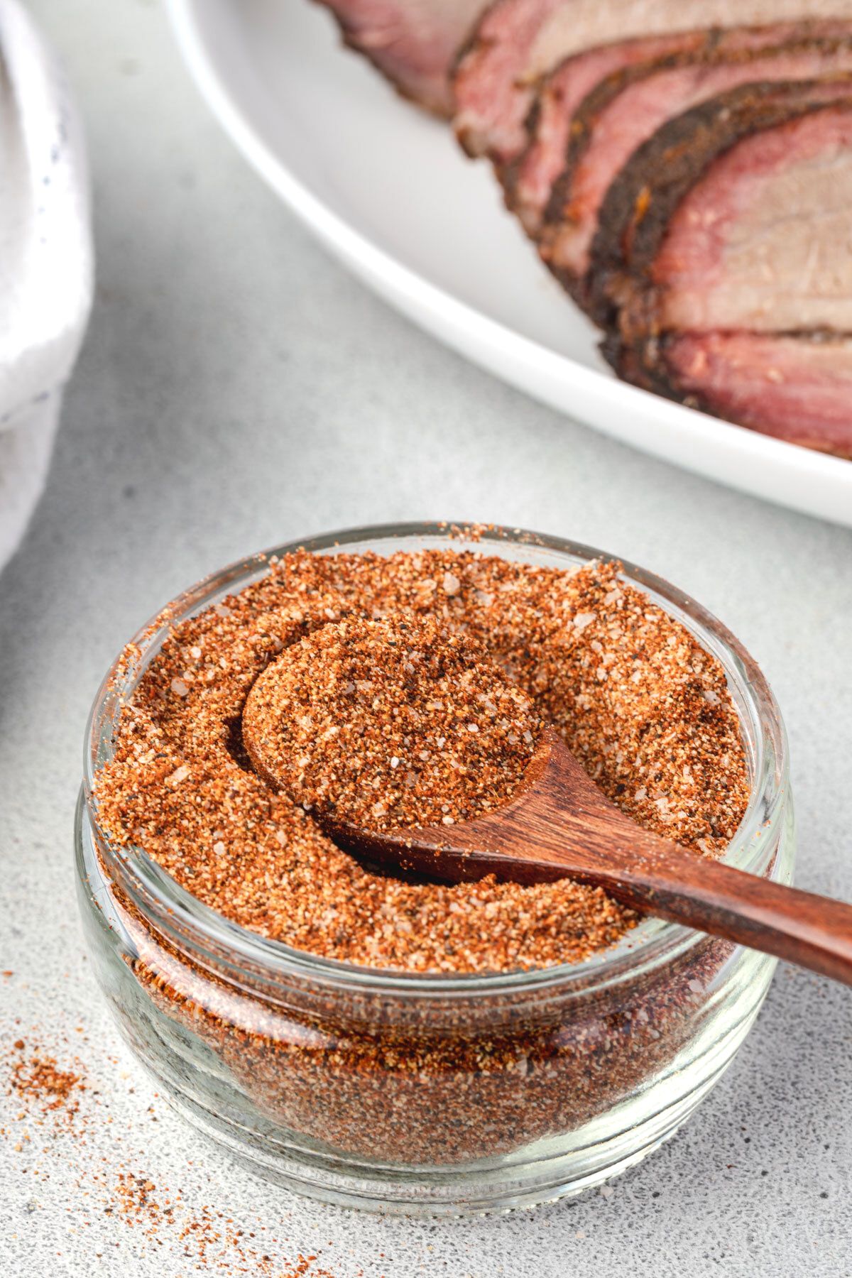 Shallow jar of spice mix with wooden spoon scooping spices and a platter of sliced brisket in background.
