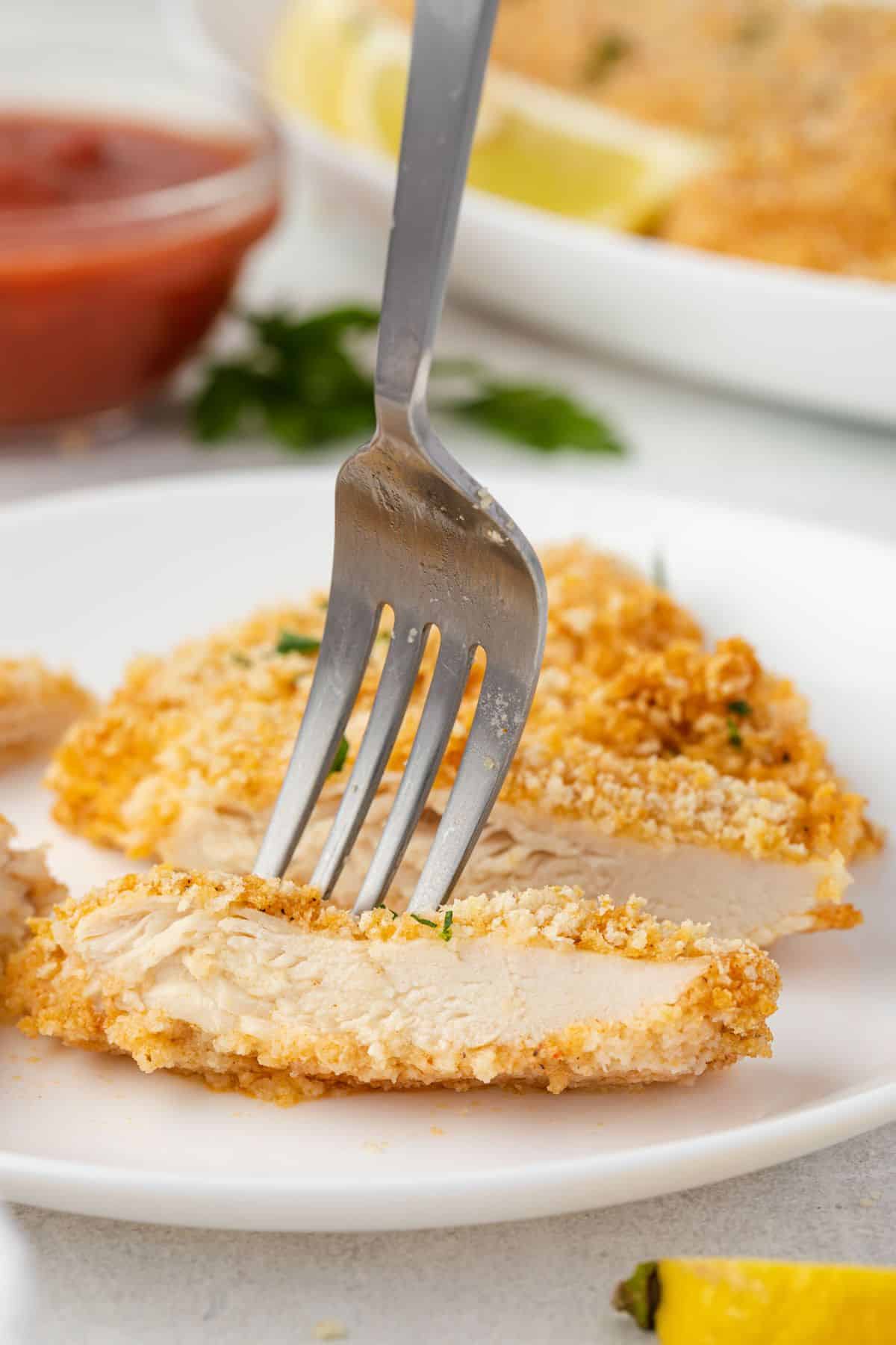 A strip of chicken on a plate with a fork sticking in it and a larger piece of chicken behind it.
