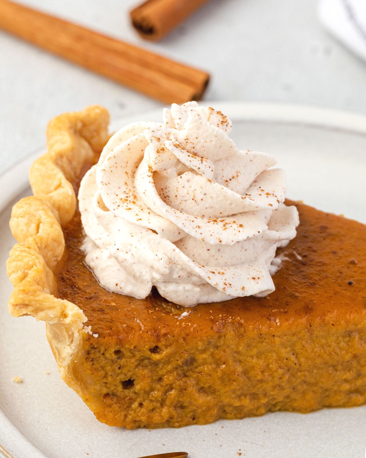 Spiced whipped cream on a slice of pumpkin pie with cinnamon sticks in background.