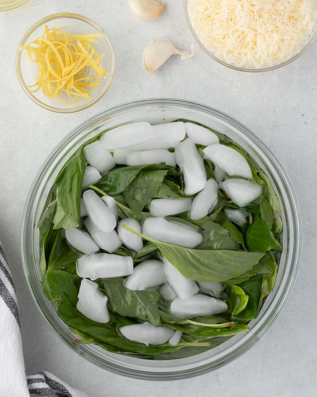 Basil leaves in a bowl of ice water with other recipe ingredients scattered around bowl.