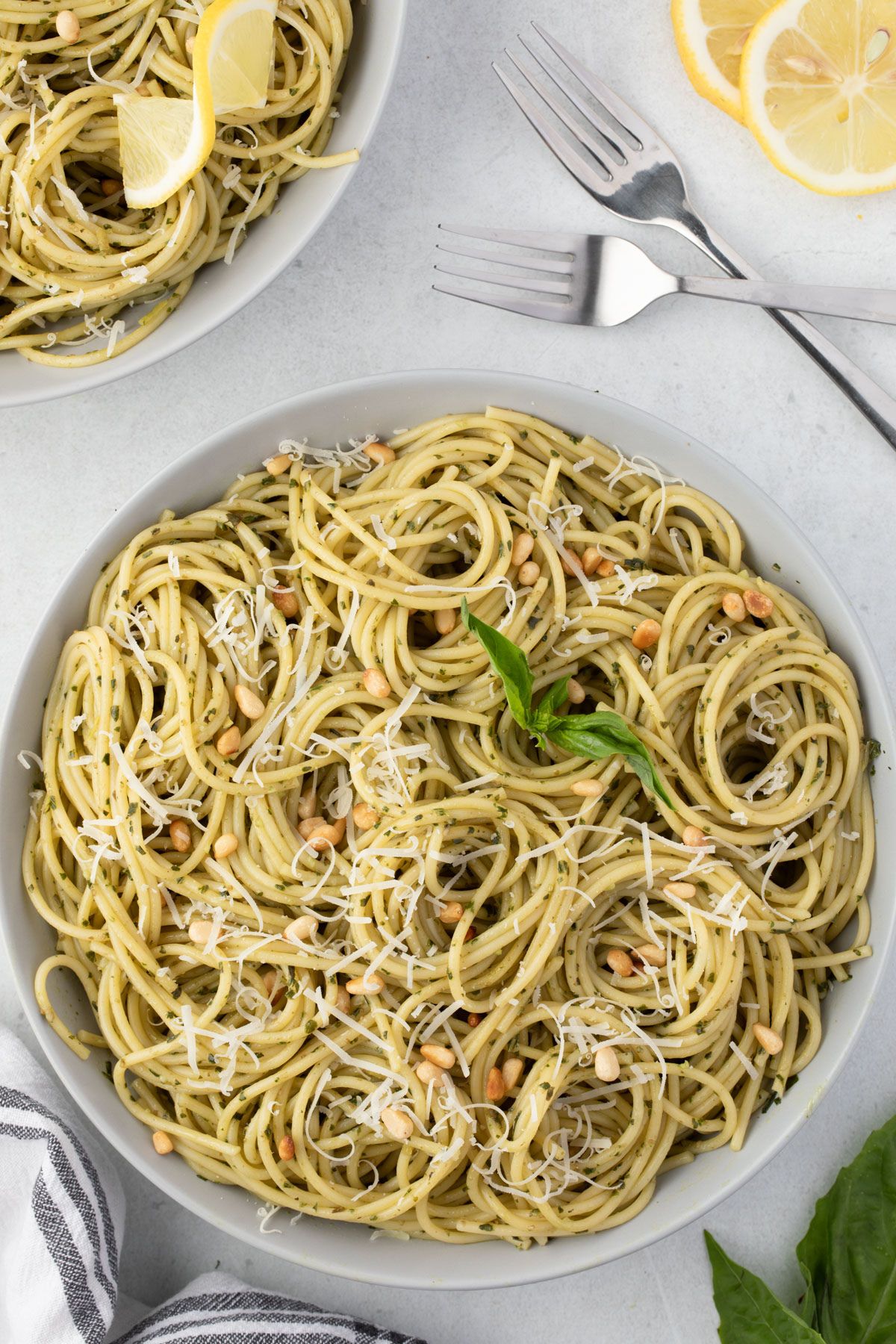 Overhead of a shallow serving bowl of pesto pasta with a second bowl, forks, and lemon slices at top of image.