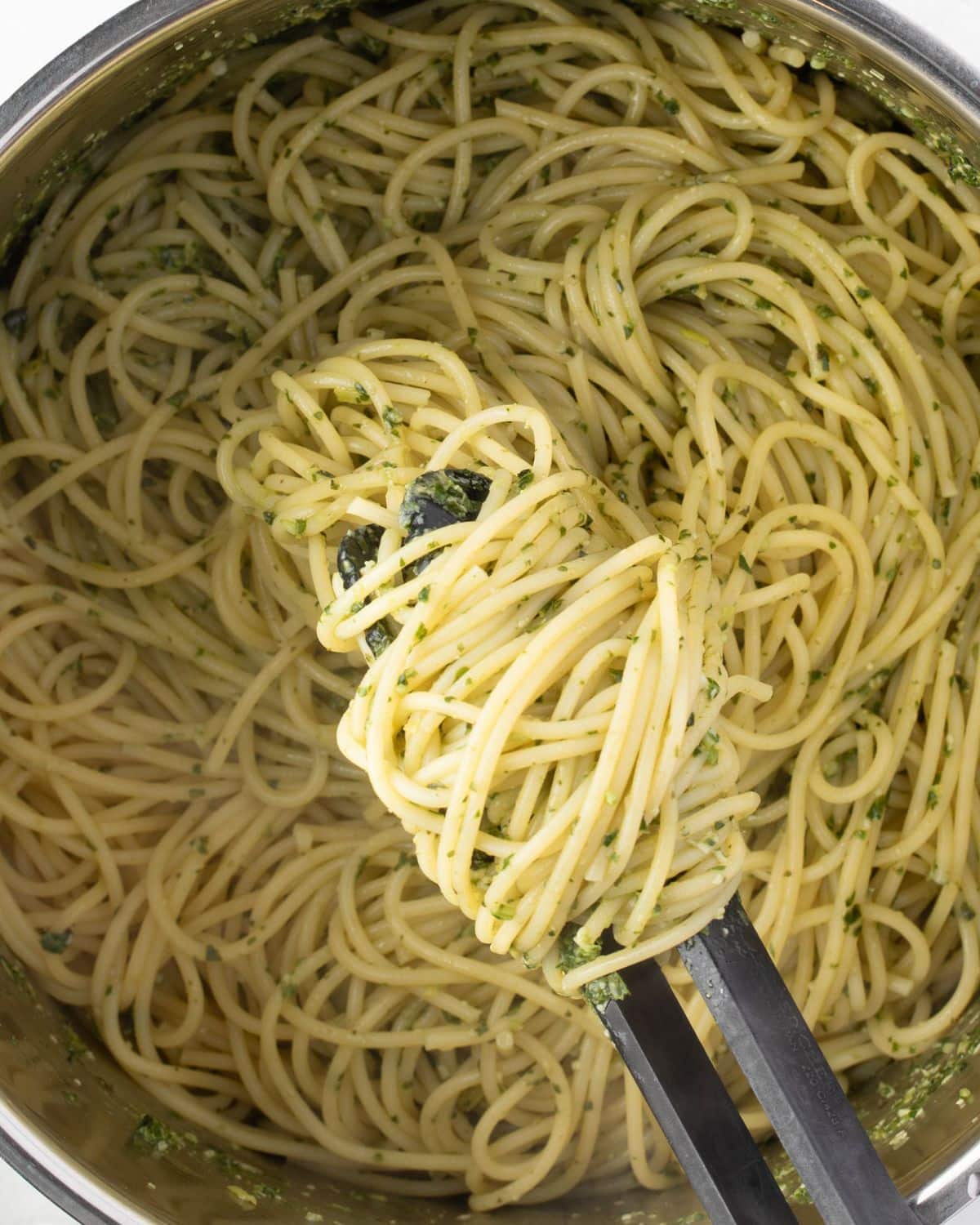 Close up of pesto spaghetti twirled around tongs, helod over a pot of noodles.