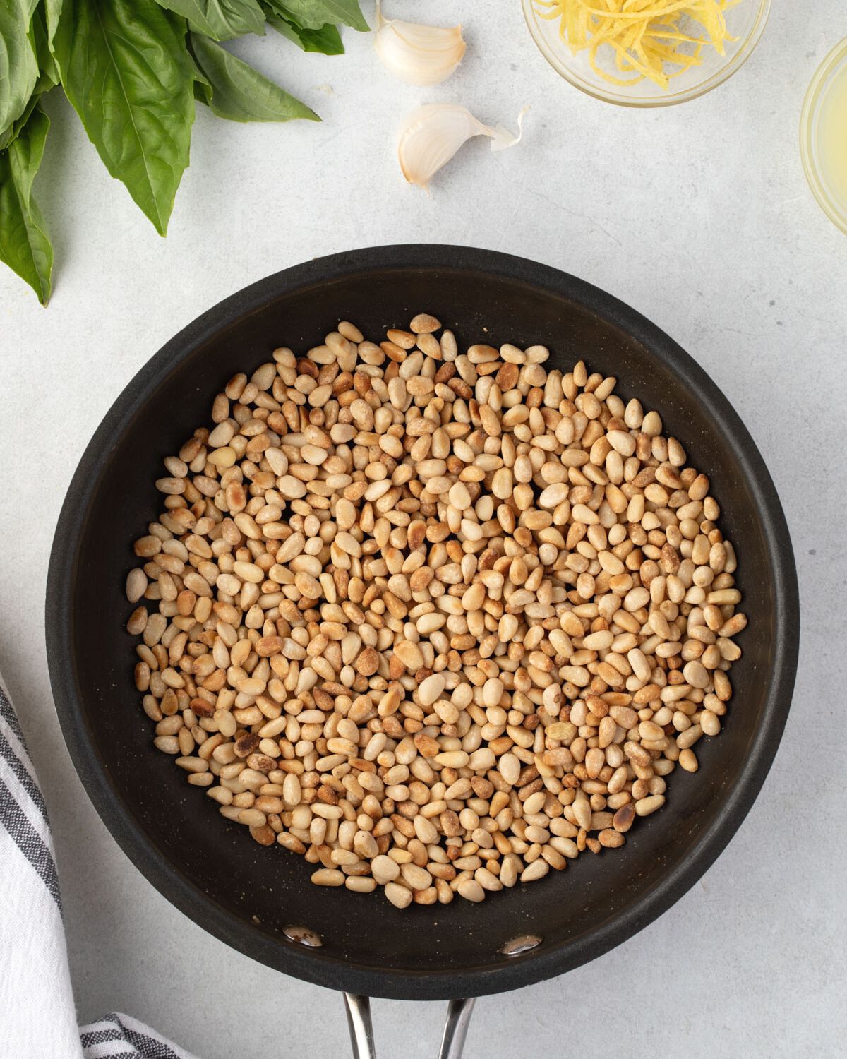 Toasted pinenuts in a skillet, with basil, garlic cloves, and lemon zest around it.