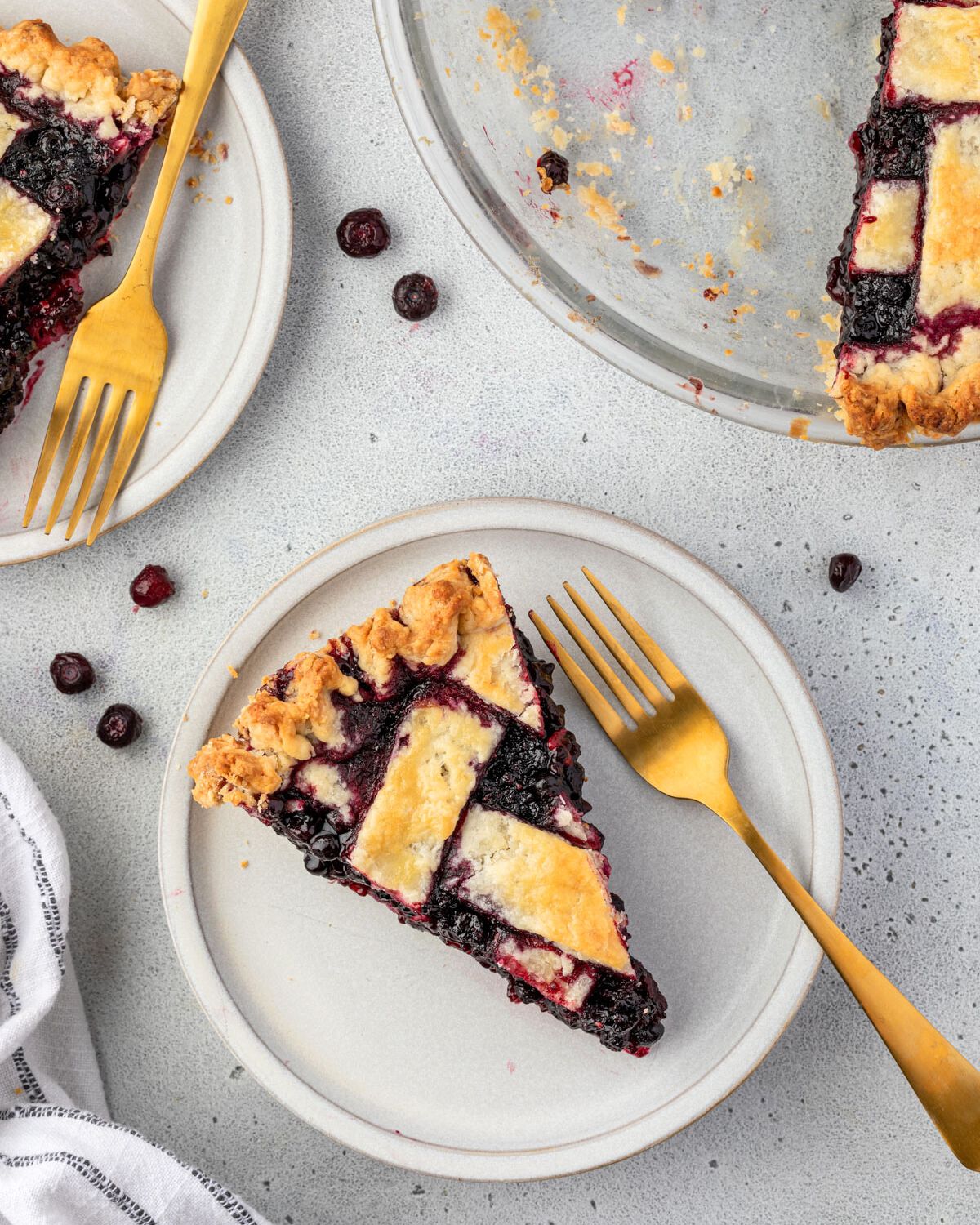 A slice of huckleberrie cake on a plate with a gold fork resting in it, another plate with pie in the corner, pie plate with some slices removed in right corner, and huckleberries sprinkled around.
