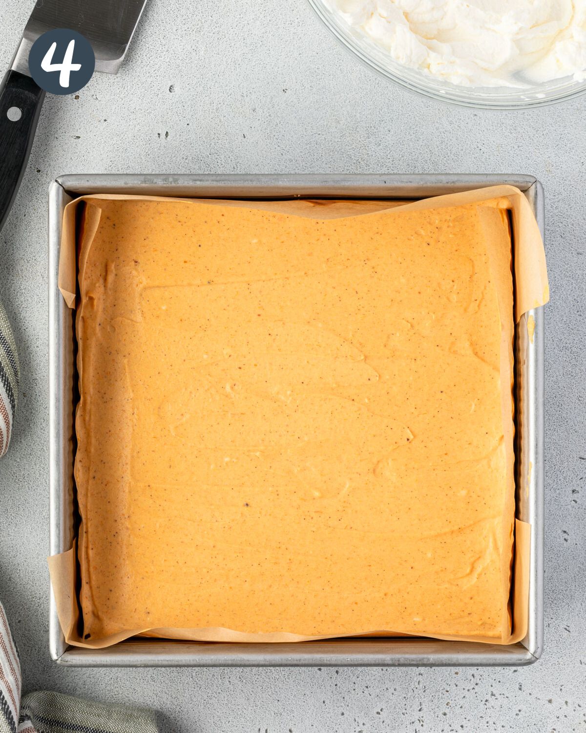 A square baking pan lined with parchment paper and pumpkin cheesecake filling spread over the top.