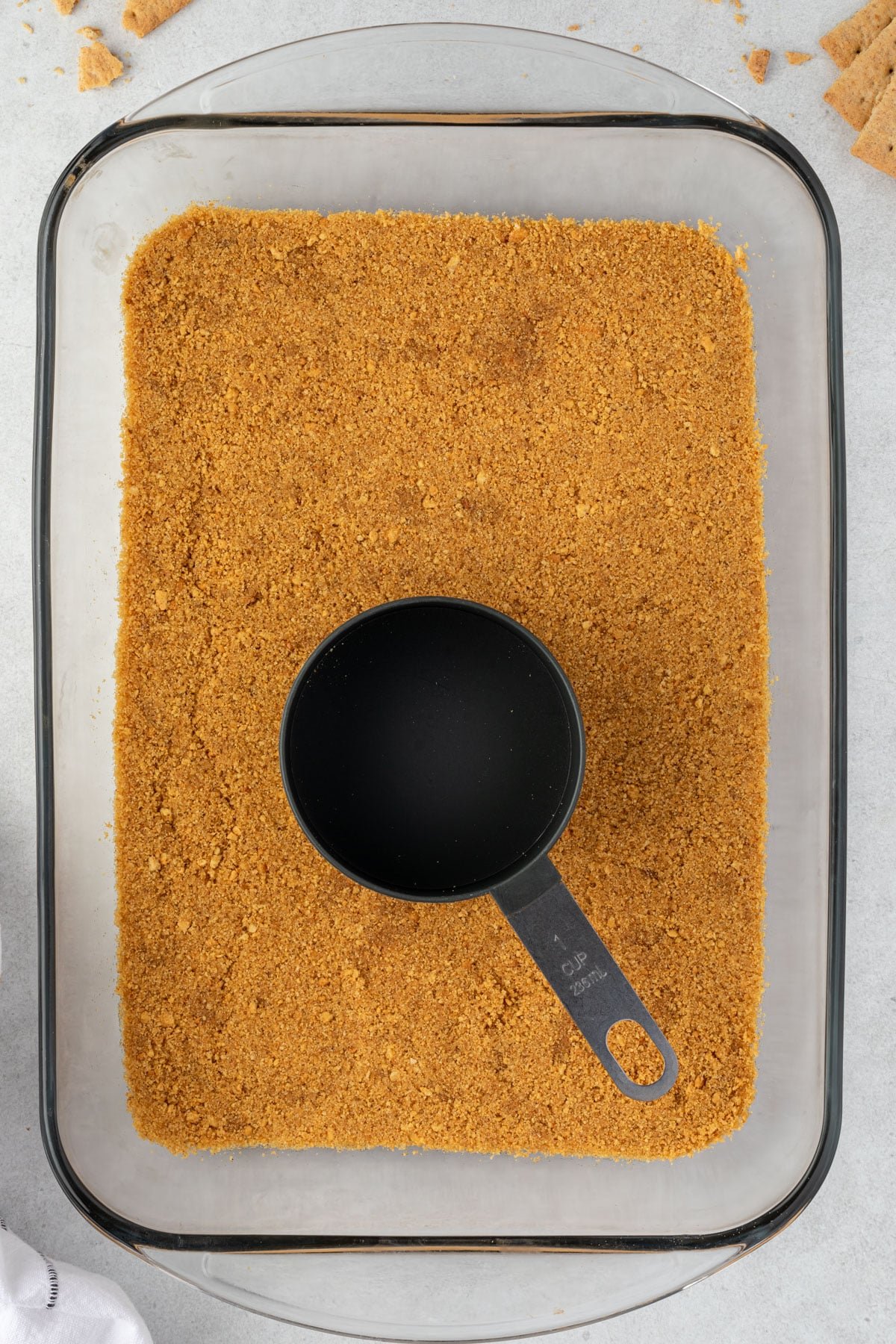 A glass 9x13 pan with a graham cracker crust pressed into it and a black measurign cup centered on the crust.