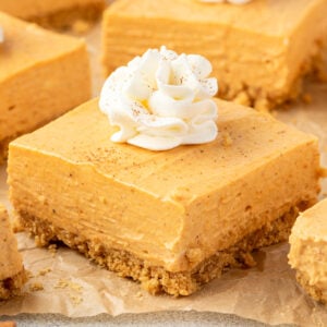 A square pumpkin cheesecake bar showing ¼-inch thick graham cracker crust, creamy filling, topped with whipped cream.