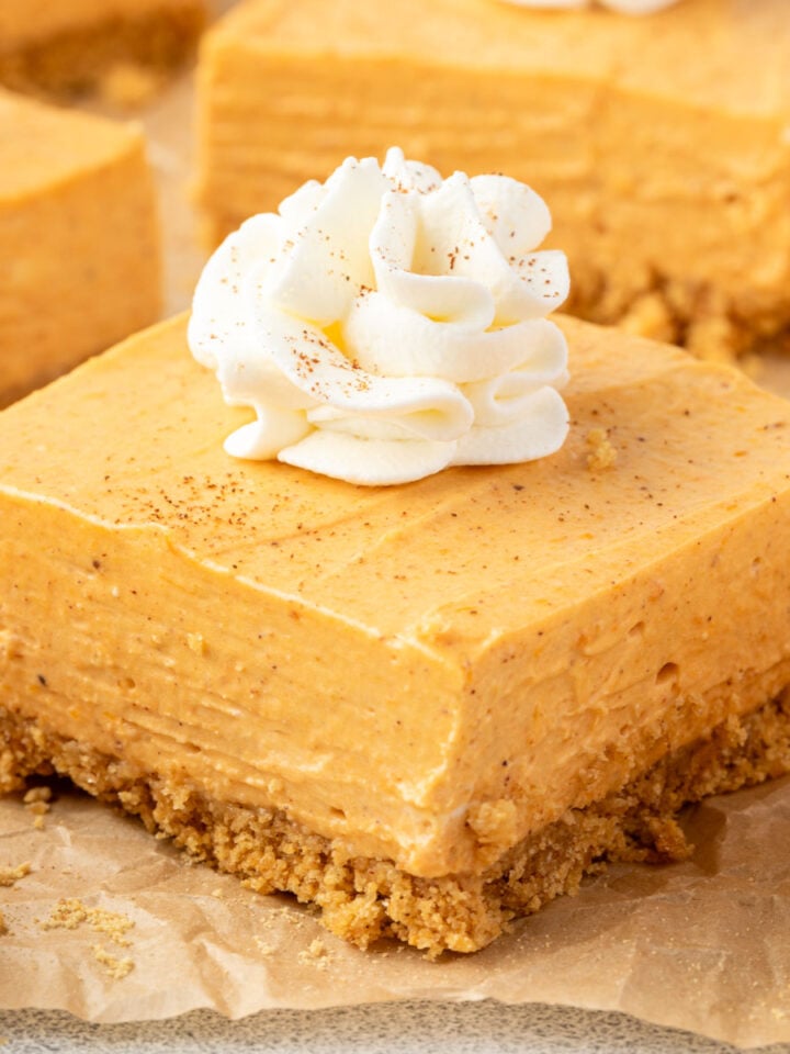 A square pumpkin cheesecake bar showing ¼-inch thick graham cracker crust, creamy filling, topped with whipped cream.