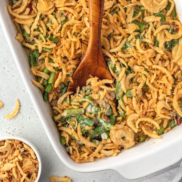 A wooden spoon digging into no mushroom green bean casserole, and a small bowl of fried onions on the side.