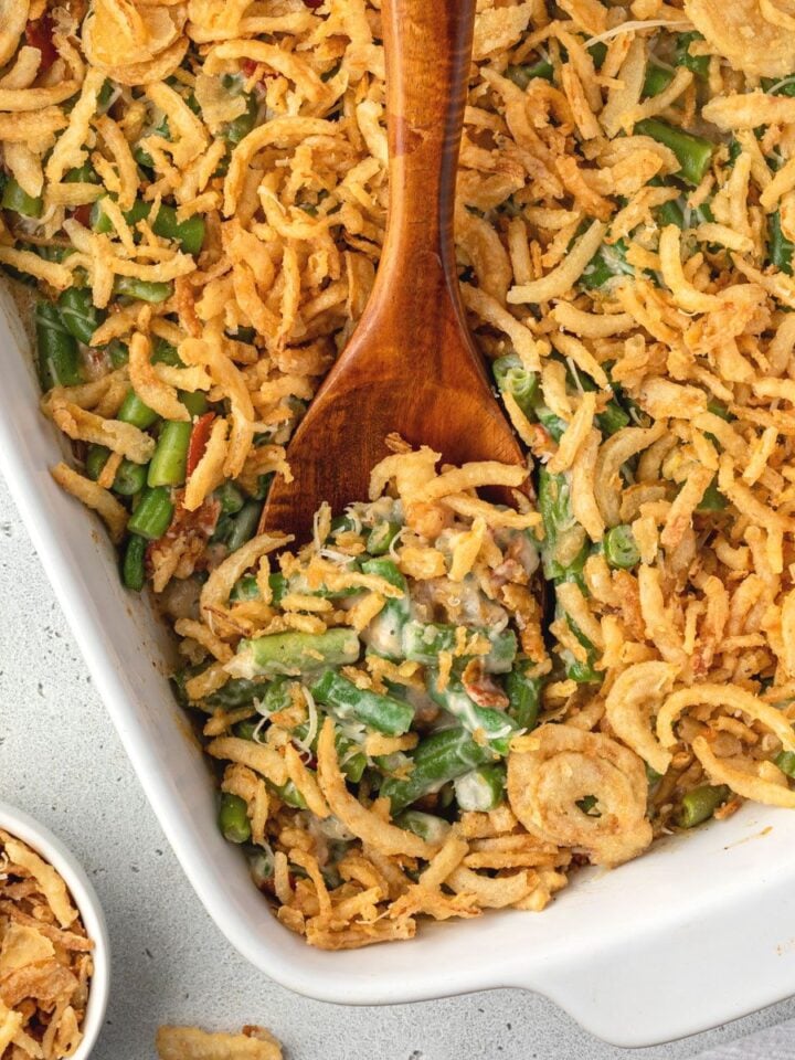 A wooden spoon digging into no mushroom green bean casserole, and a small bowl of fried onions on the side.