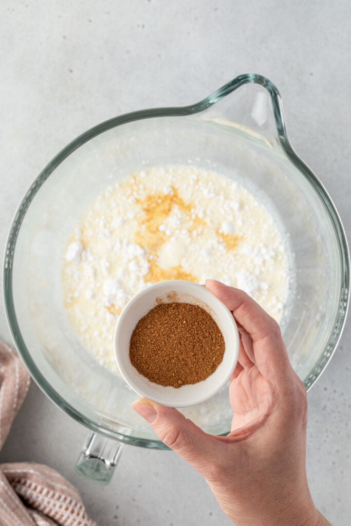 A hand holding a small white bowl of pumpkin spice over a large mixing bowl of cream.