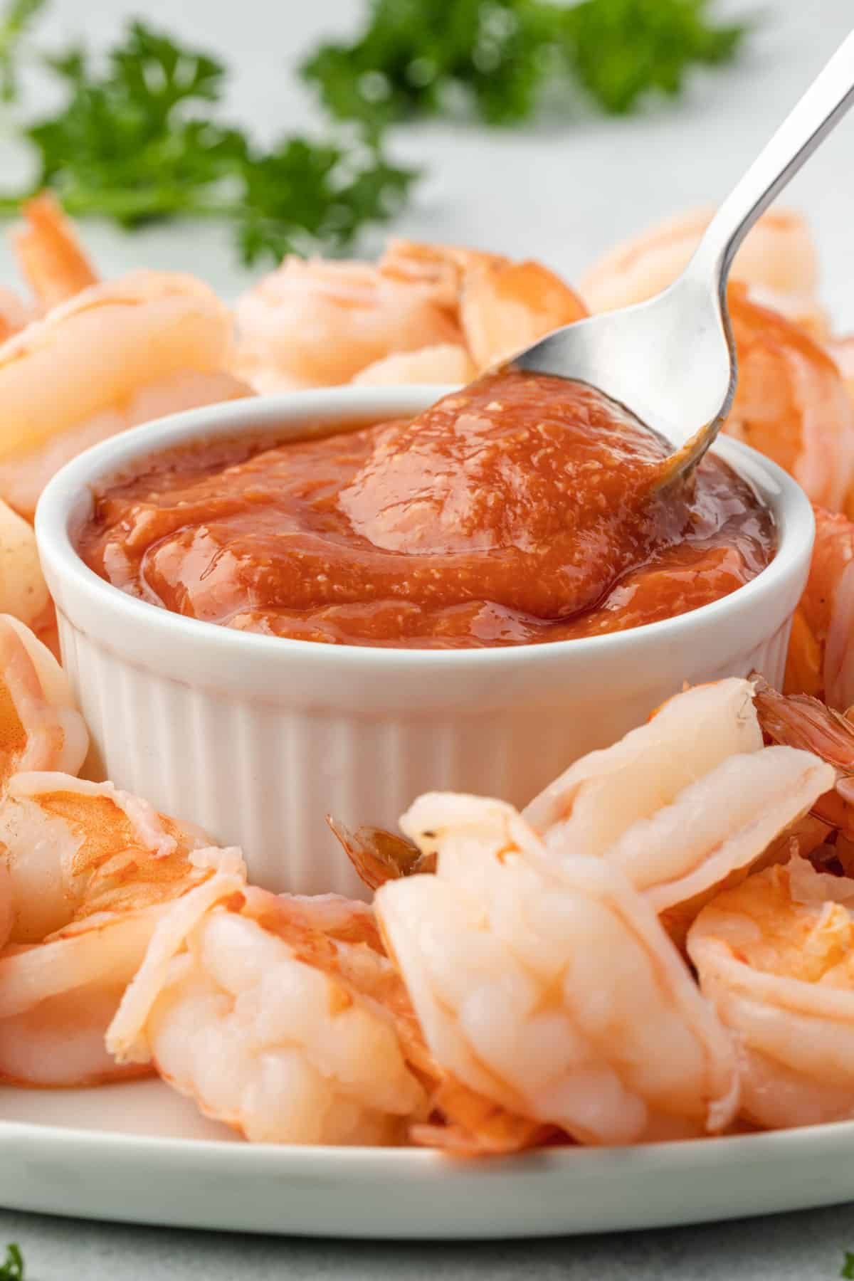 A white bowl filled with cocktail sauce that has a spoon scooping some sauce out, on a platter of shrimp.
