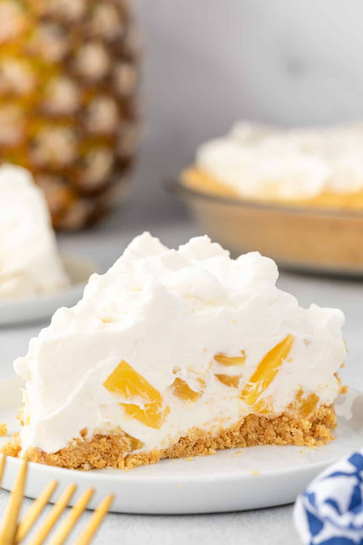 A slice of pineapple cream cheese pie on a plate with a pineapple and the pie in the background.