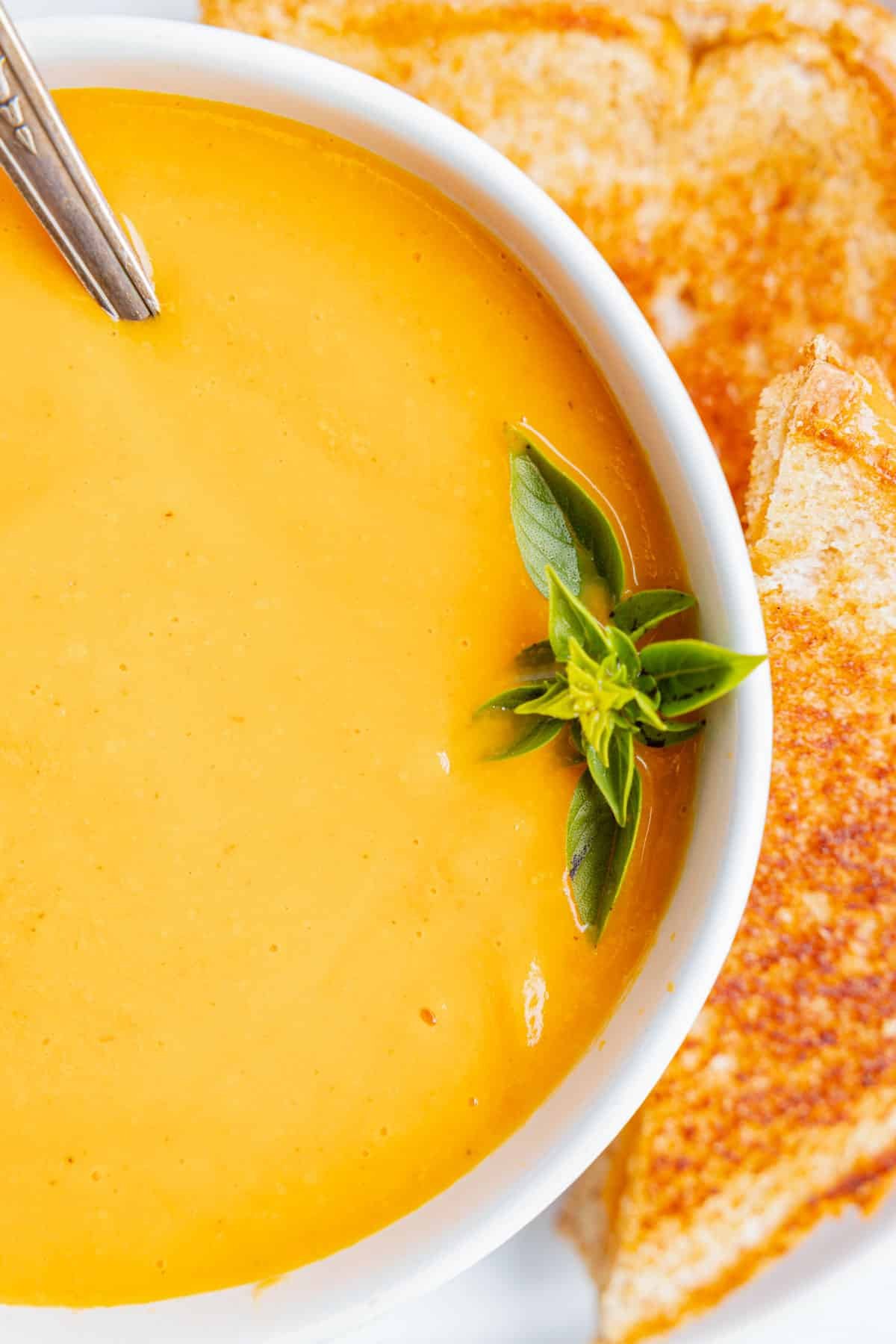 A bowl of roasted butternut squash and red pepper soup in a bowl with a sprig of basil and grilled cheese sandwiches wedge in on a plate.