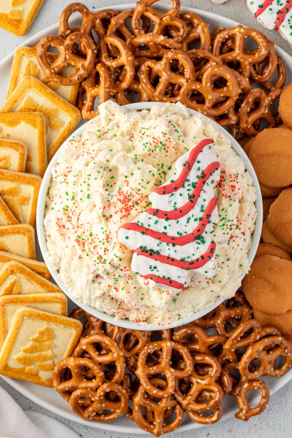 Cream cheese dip with red and green sanded sugar sprinkles in a white bowl, garnished with a Christmas tree cake on a platter with pretzels and cookies.