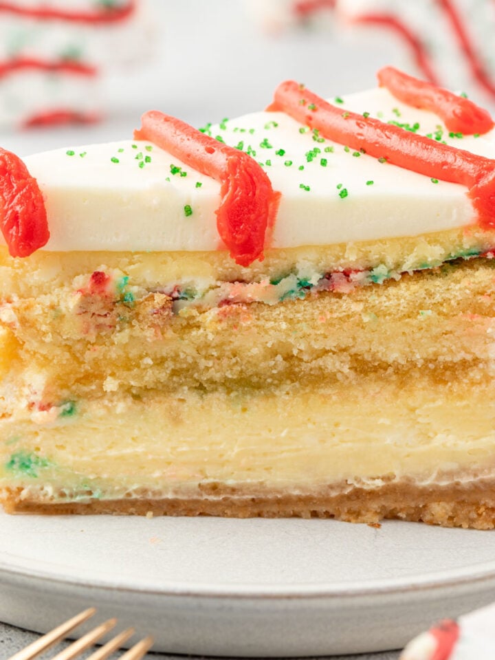 Slice of cheesecake on a plate, showing layers of crust, cheesecake, cake, more cheesecake, and frosting, with Little Debbie trees standing in background.