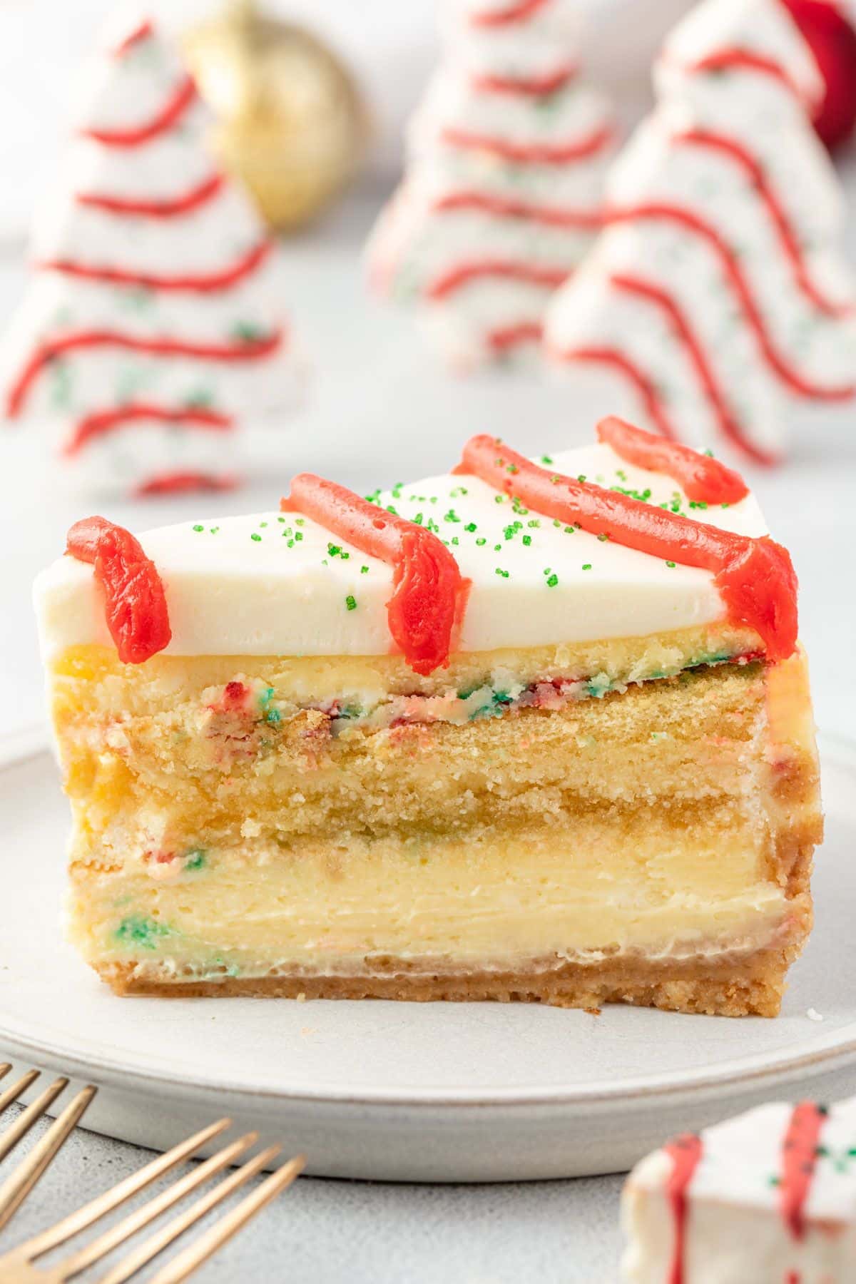Slice of cheesecake on a plate, showing layers of crust, cheesecake, cake, more cheesecake, and frosting, with Little Debbie trees standing in background.